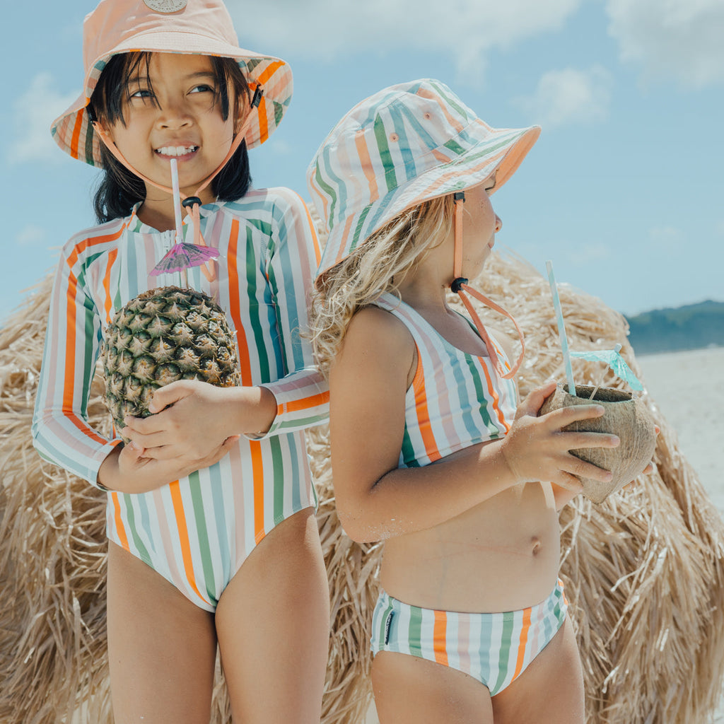 Two young children wearing the CRYWOLF bikini and long sleeved swimsuit while enjoying a drink on the beach.