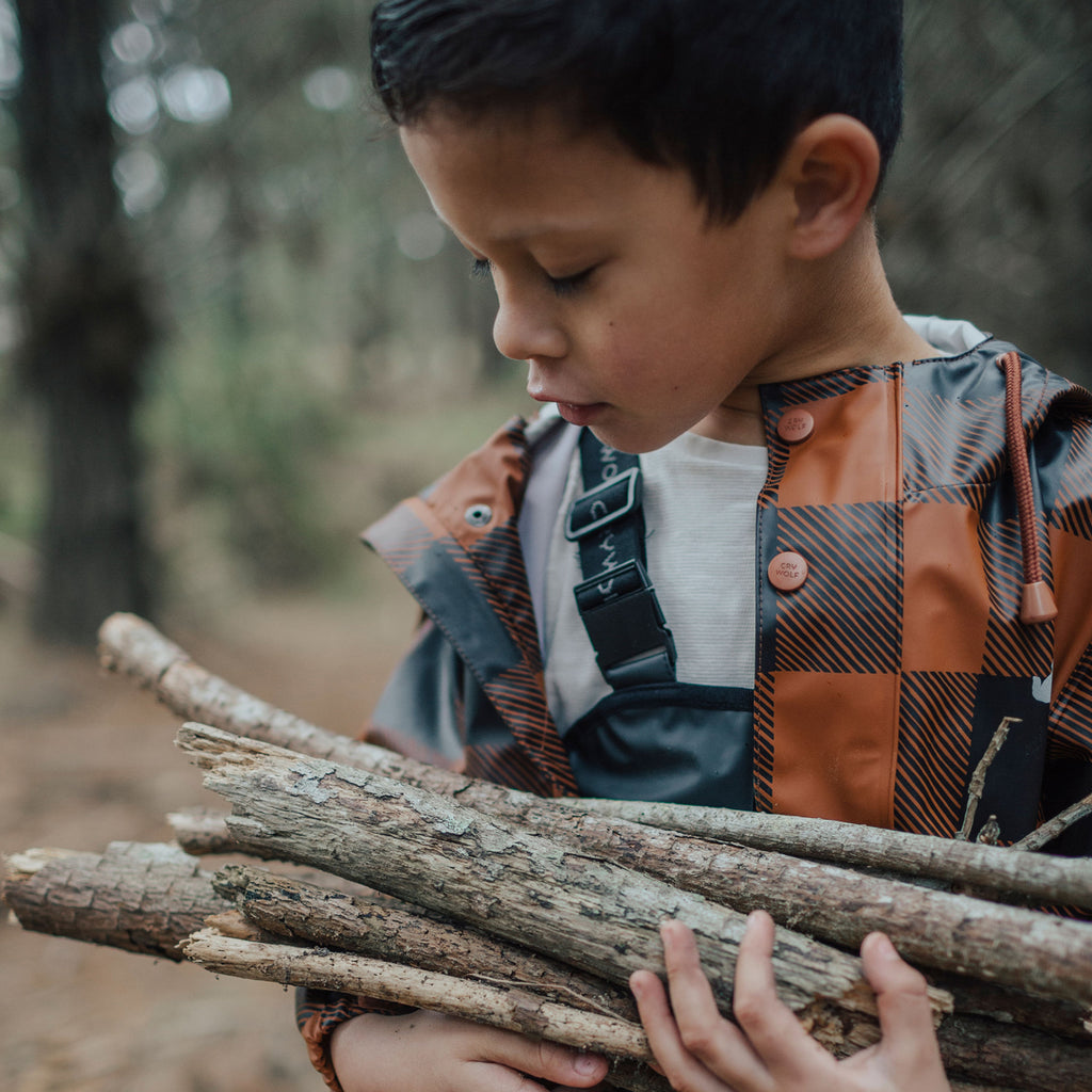 A boy carrying logs in a forest wearing a Crywolf Play Jacket Rust Plaid