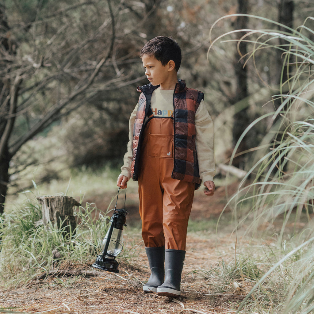 A Boy Walking Through A Forest Holding A Lantern Wearing Crywolf Rain Overalls Rust Made From Recycled Fabrics - Front View